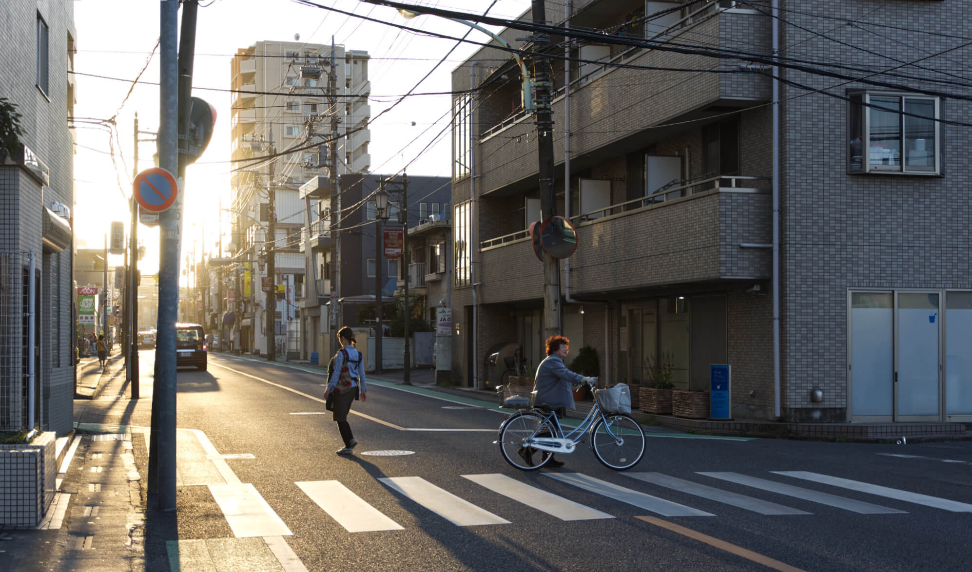 A road crossing at sunset. A young woman crosses the street to the light, her shadow stretching outwards. An older woman wheels a bike towards the opposite side. A mess of cables criss-crosses overhead.