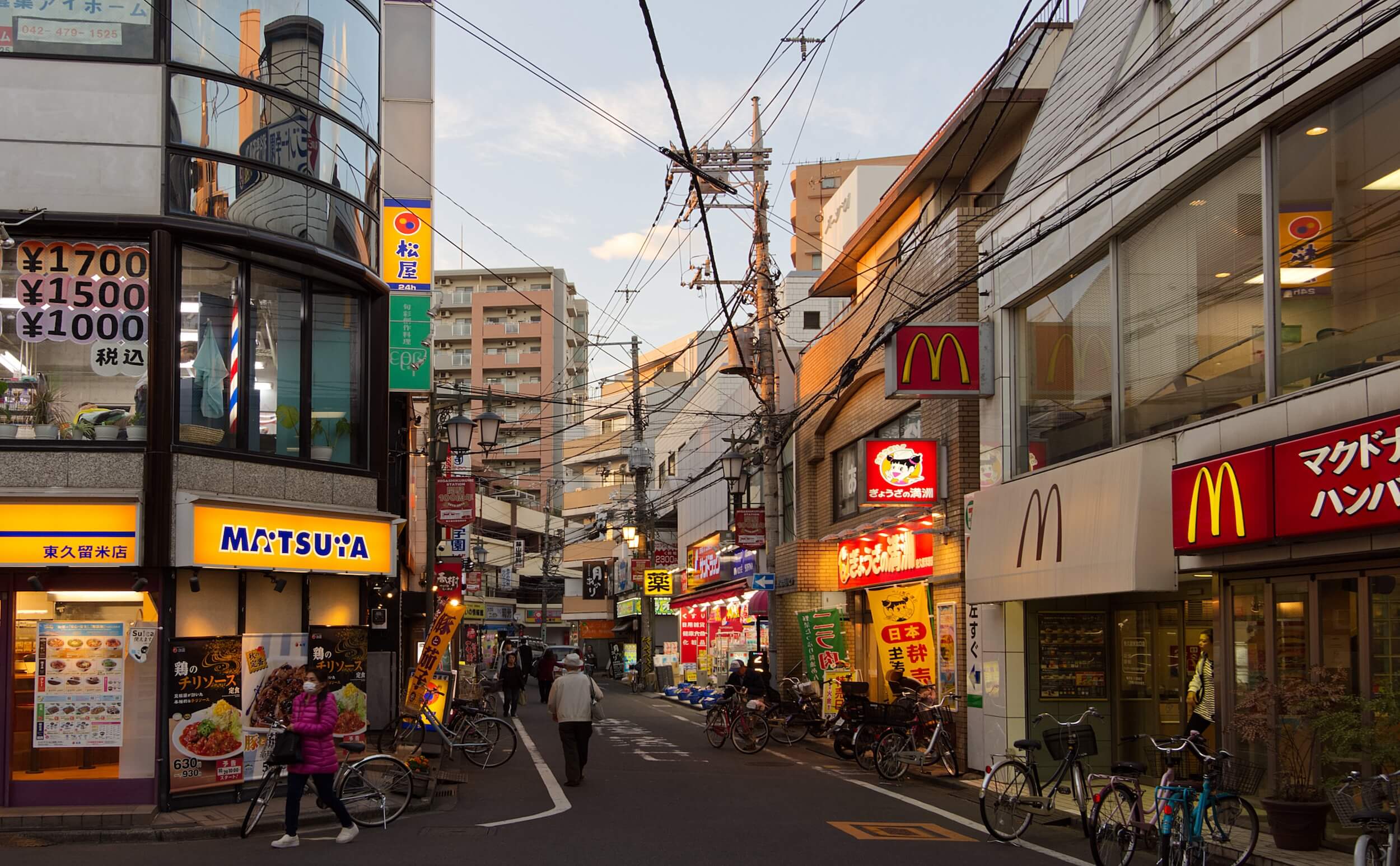 A street at sunset. In the foreground a Matsuya restaurant is illuminated. On the opposite side of the street is a McDonalds. An older man walks away down the middle of the road