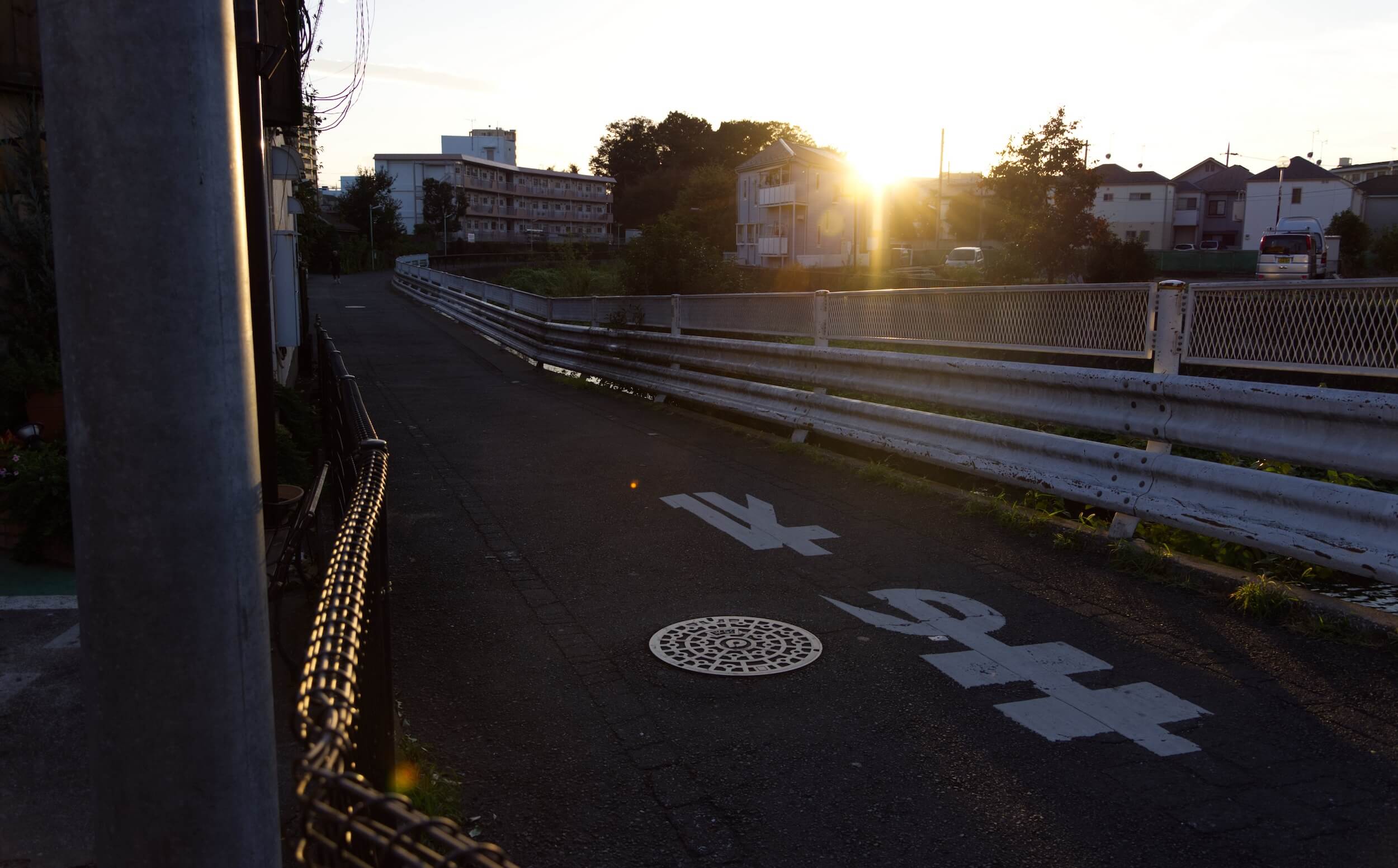 A road runs alongside a river at sunset. The street is in shadow and the light catches the edges of fences and railings